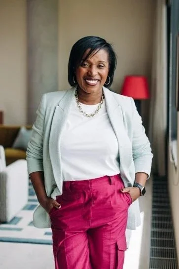 A woman smiling in a professional setting, wearing a white top, gray blazer, and pink pants.