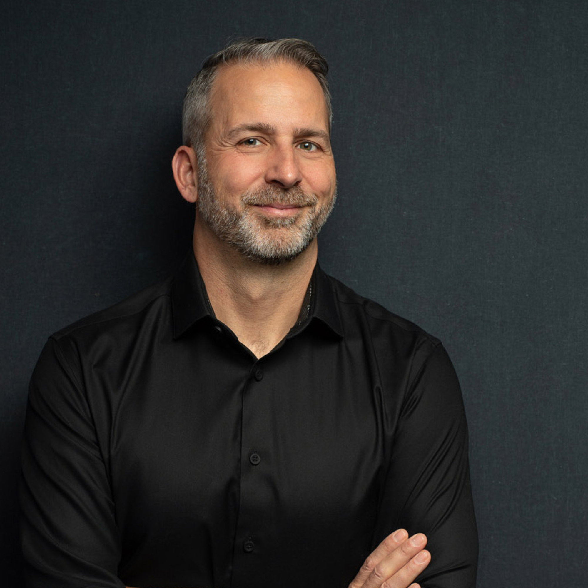 Close-up of a smiling middle-aged man with a short beard and mustache, wearing a black shirt, standing against a dark background, with his arms crossed.