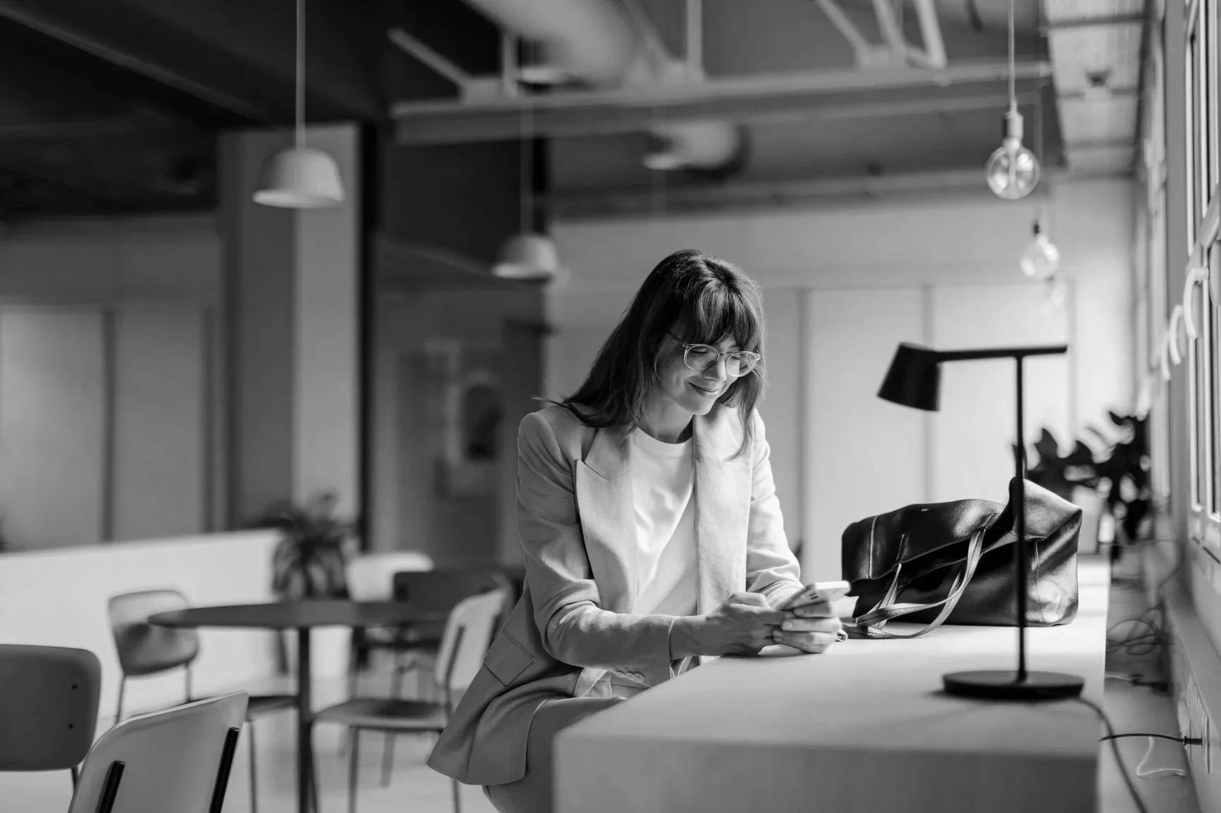 A woman with glasses and shoulder-length hair sits at a table in a modern office, looking at her phone with a smile. She is dressed in a light blazer and shirt. There is a bag and a desk lamp on the table, and the office has hanging lights and large windows.