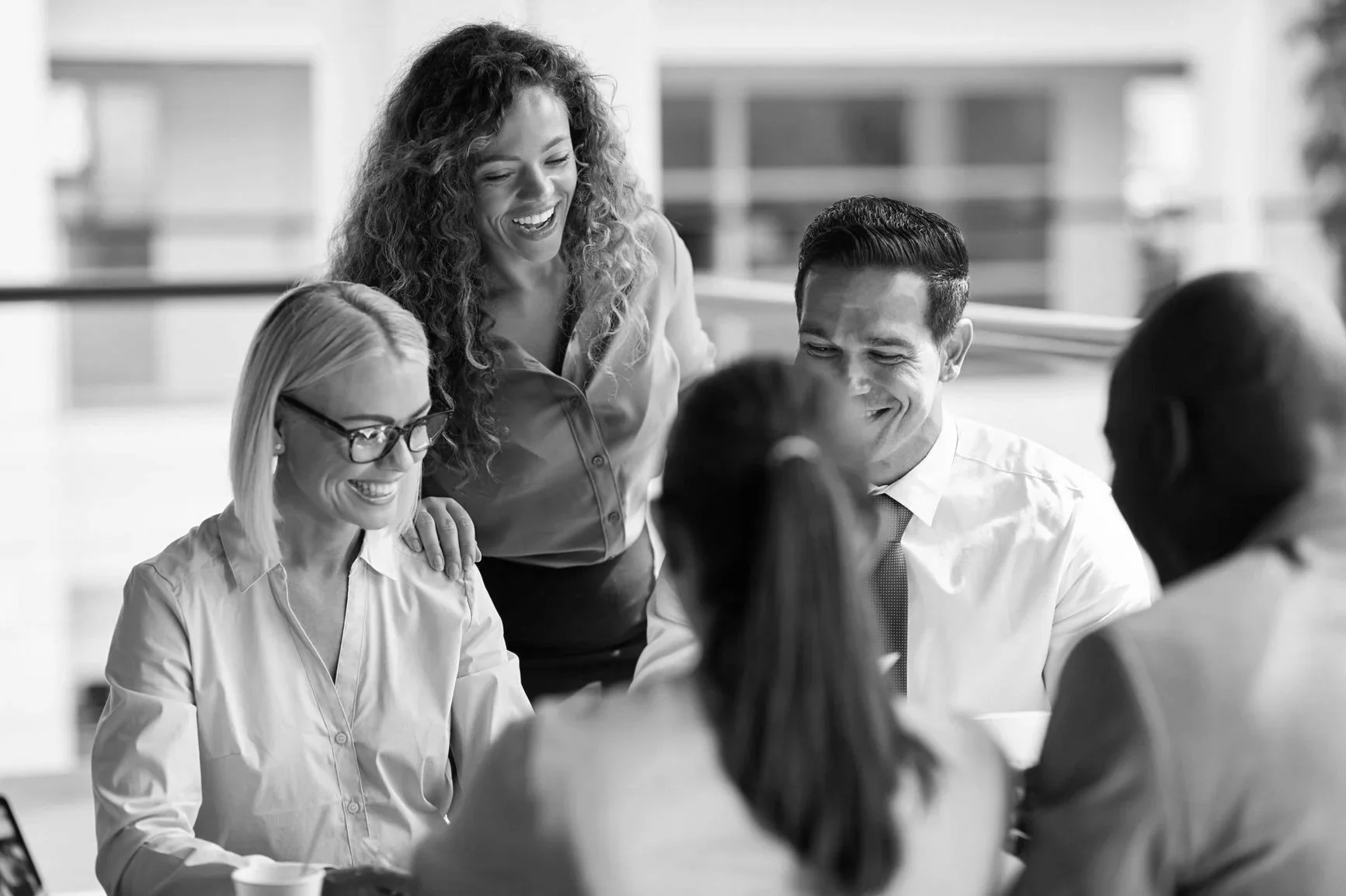 Group of five diverse professionals in a meeting, smiling and laughing, engaged in conversation in an office setting.