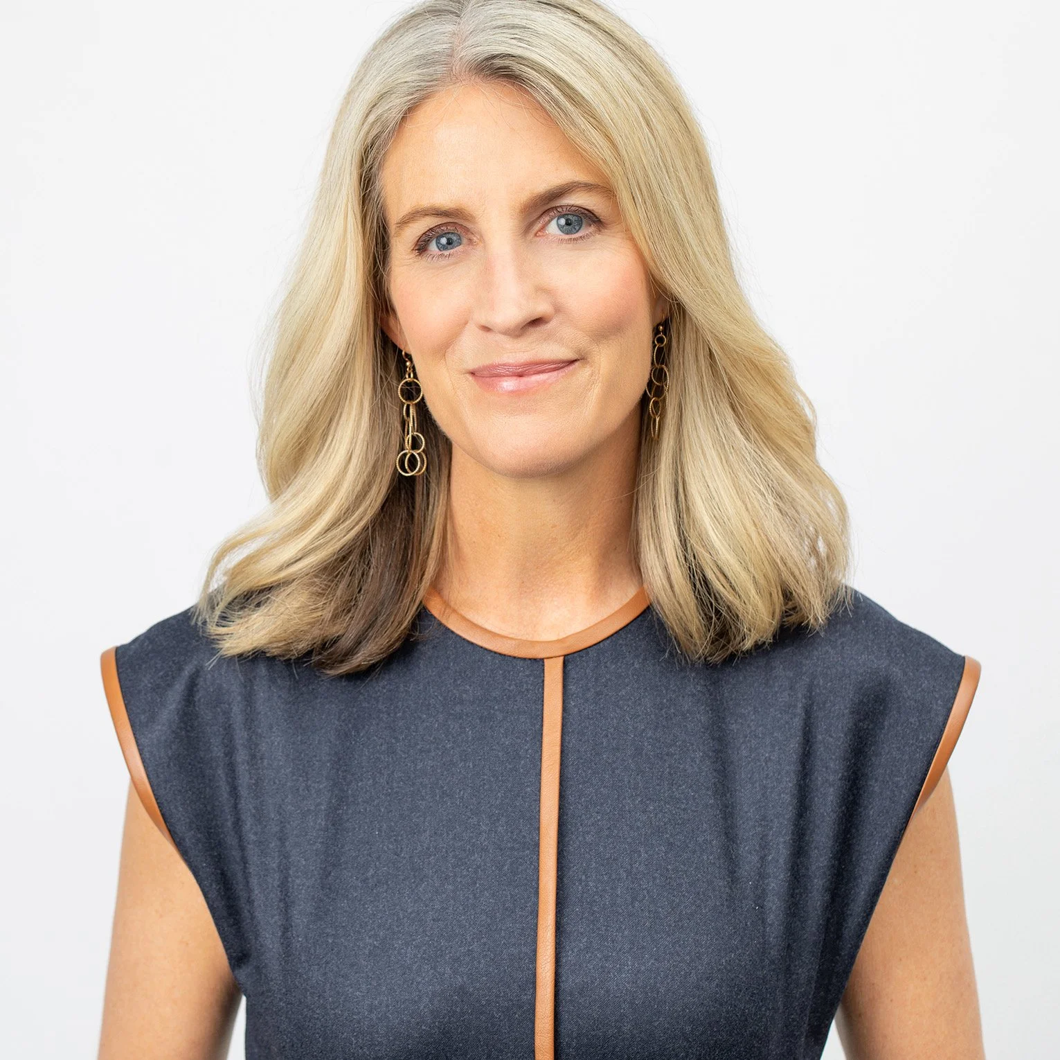 A portrait of a middle-aged woman with shoulder-length blonde hair, blue eyes, wearing gold earrings, and a sleeveless navy dress with a brown trim, posing against a plain white background.