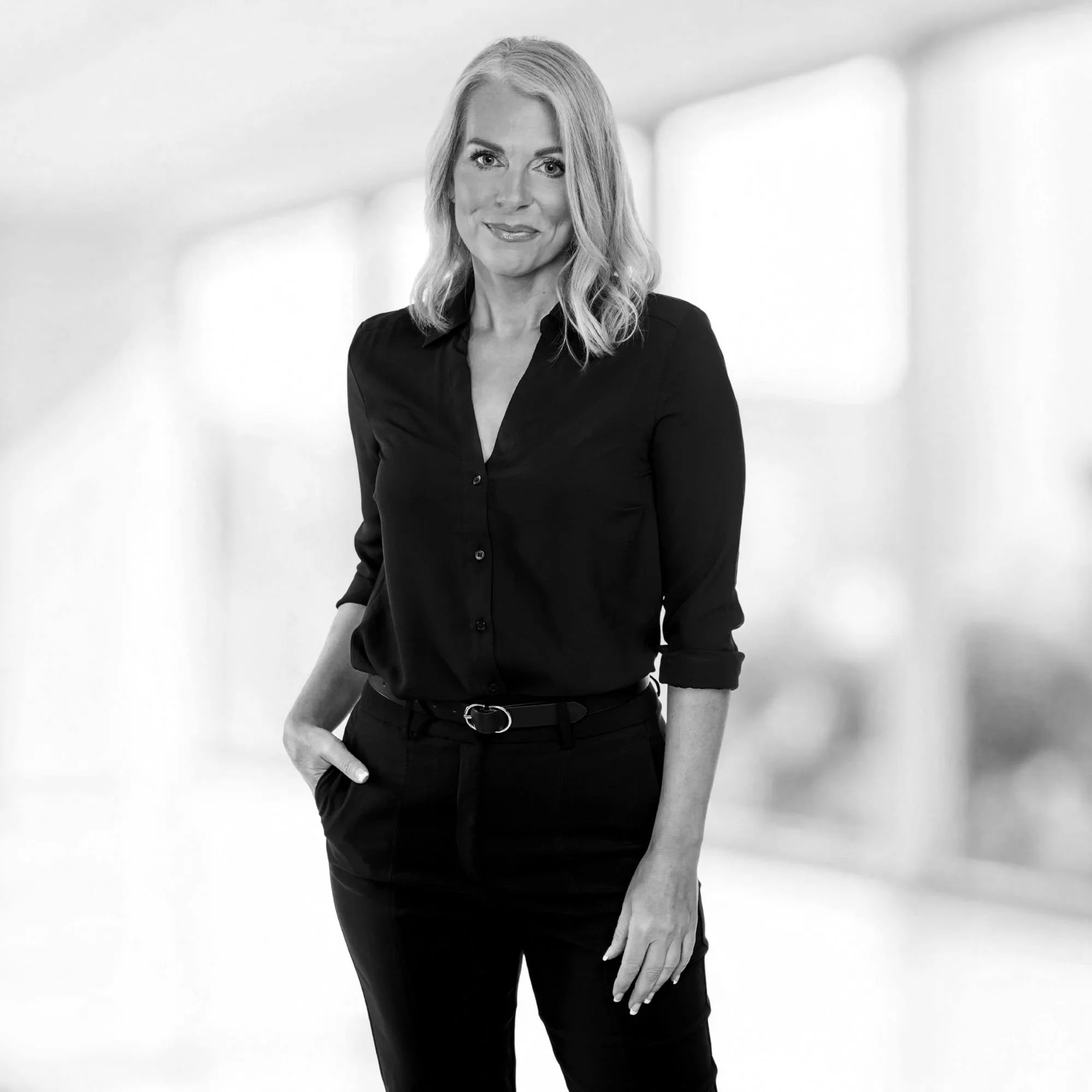 Black and white portrait of a woman with wavy blonde hair, wearing a black button-up shirt and black pants, standing in a bright, minimalistic indoor setting with large windows.