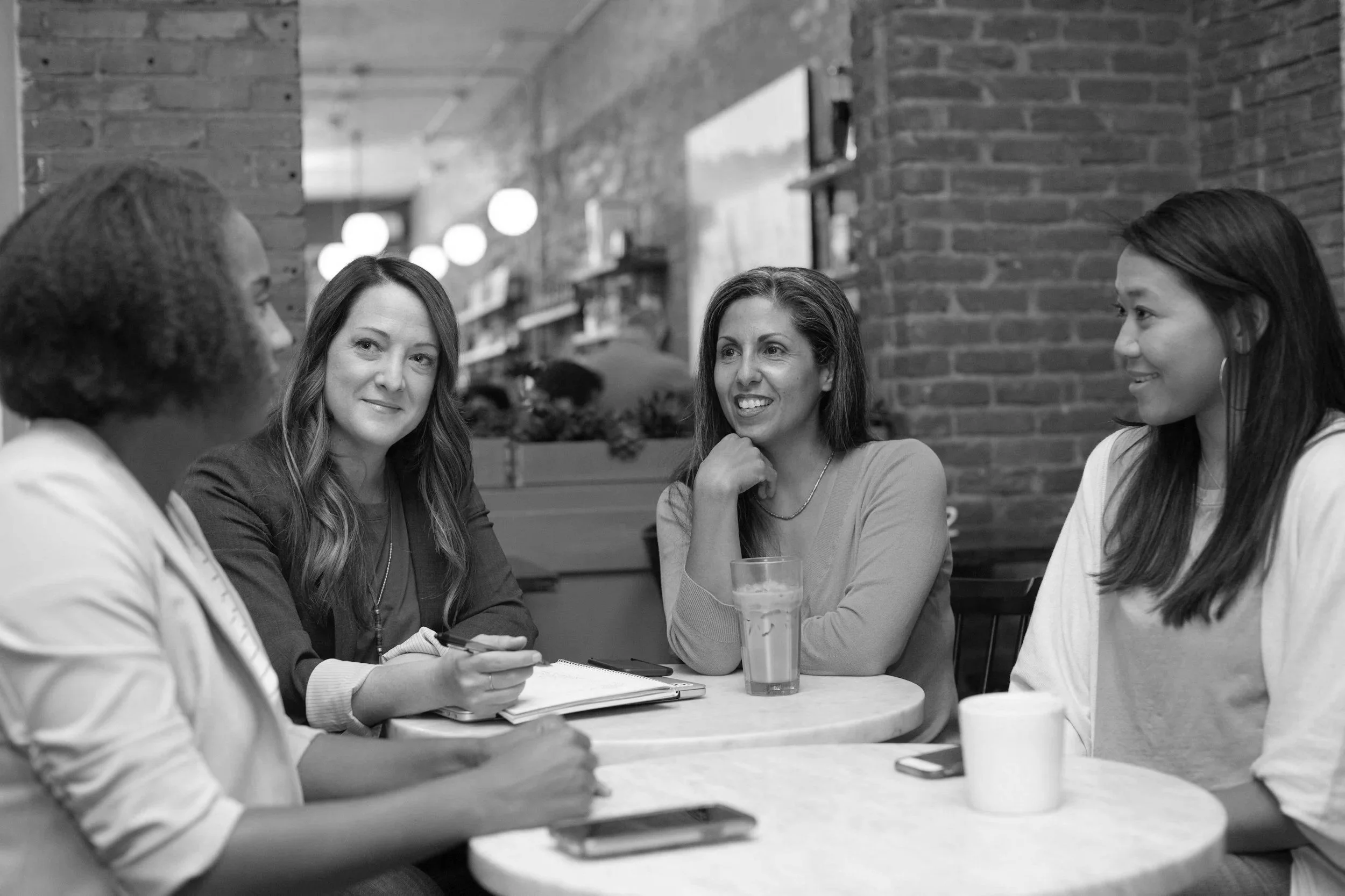 A group of women sitting around a table having a conversation in a cozy restaurant or cafe with brick walls and soft lighting.