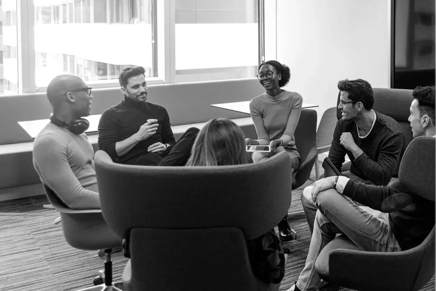 A diverse group of six people in a modern office meeting room sitting in chairs and discussing, smiling and engaging with each other.