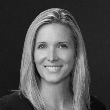 Black and white portrait of a smiling woman with straight blonde hair and light eyes, wearing a dark top, against a dark background.