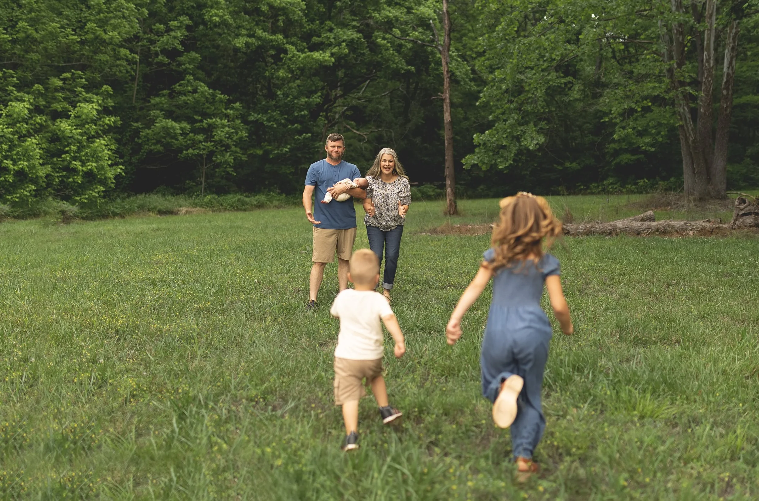 A family of five, including parents and three children, walking and running on a grassy field in a wooded area during daytime. The father is holding a baby, and the children are playing in the foreground.