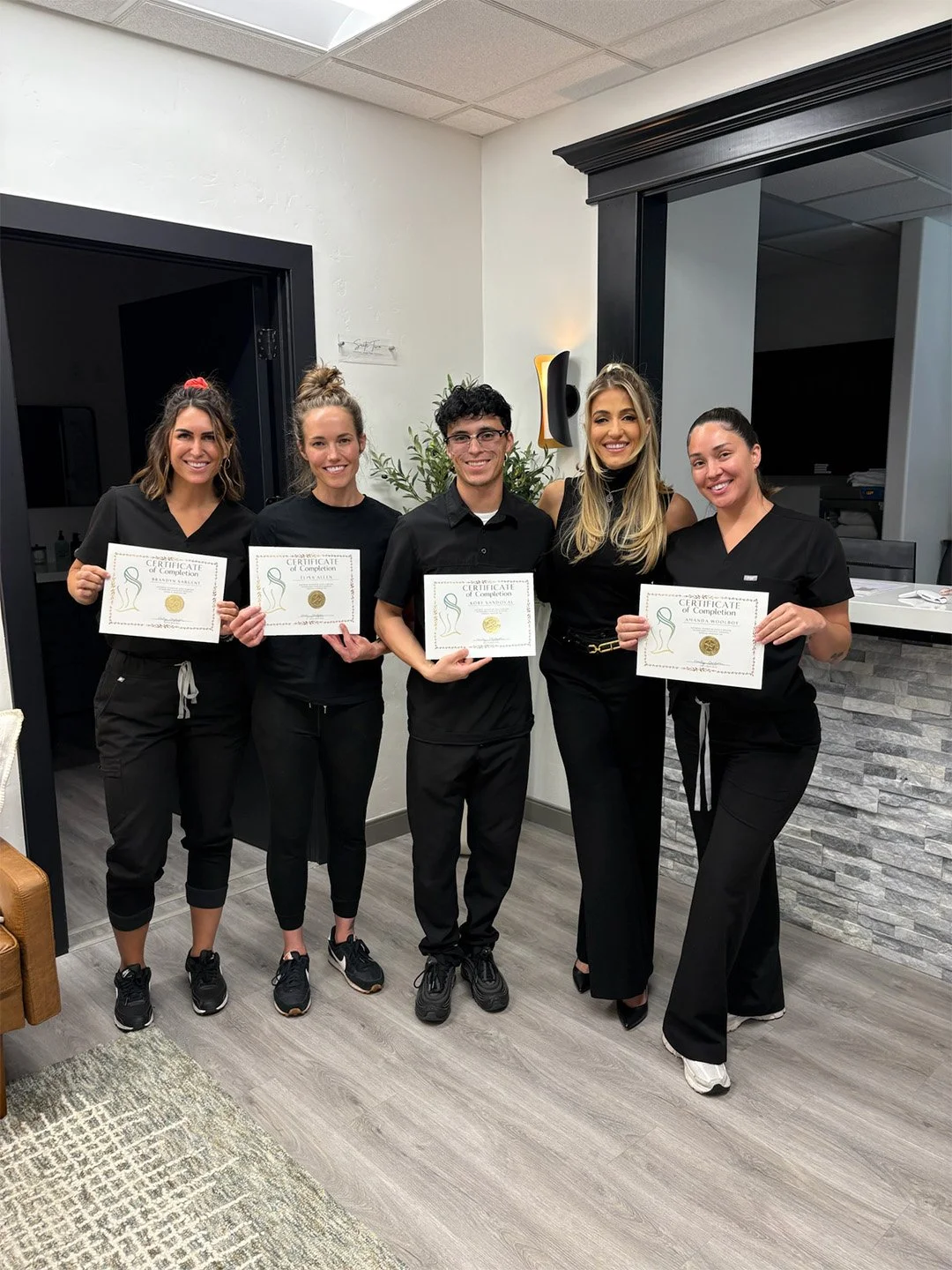 Five young adults dressed in black clothing standing indoors, holding certificates of completion, smiling at the camera, with a plant and reception area in the background.