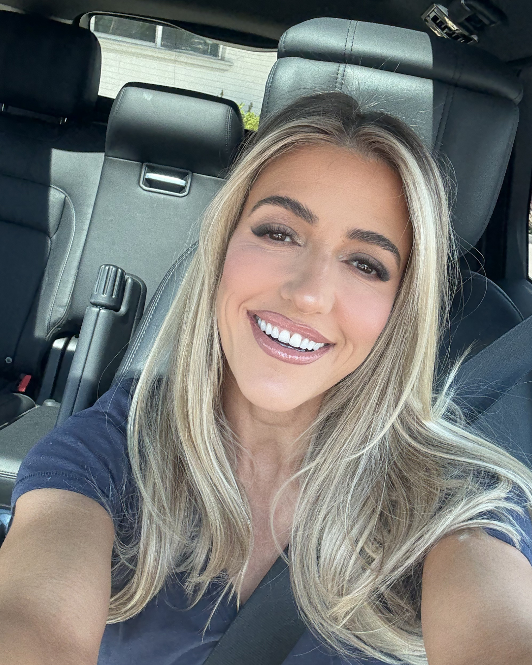 A woman with long blonde hair smiling inside a car, taking a selfie.