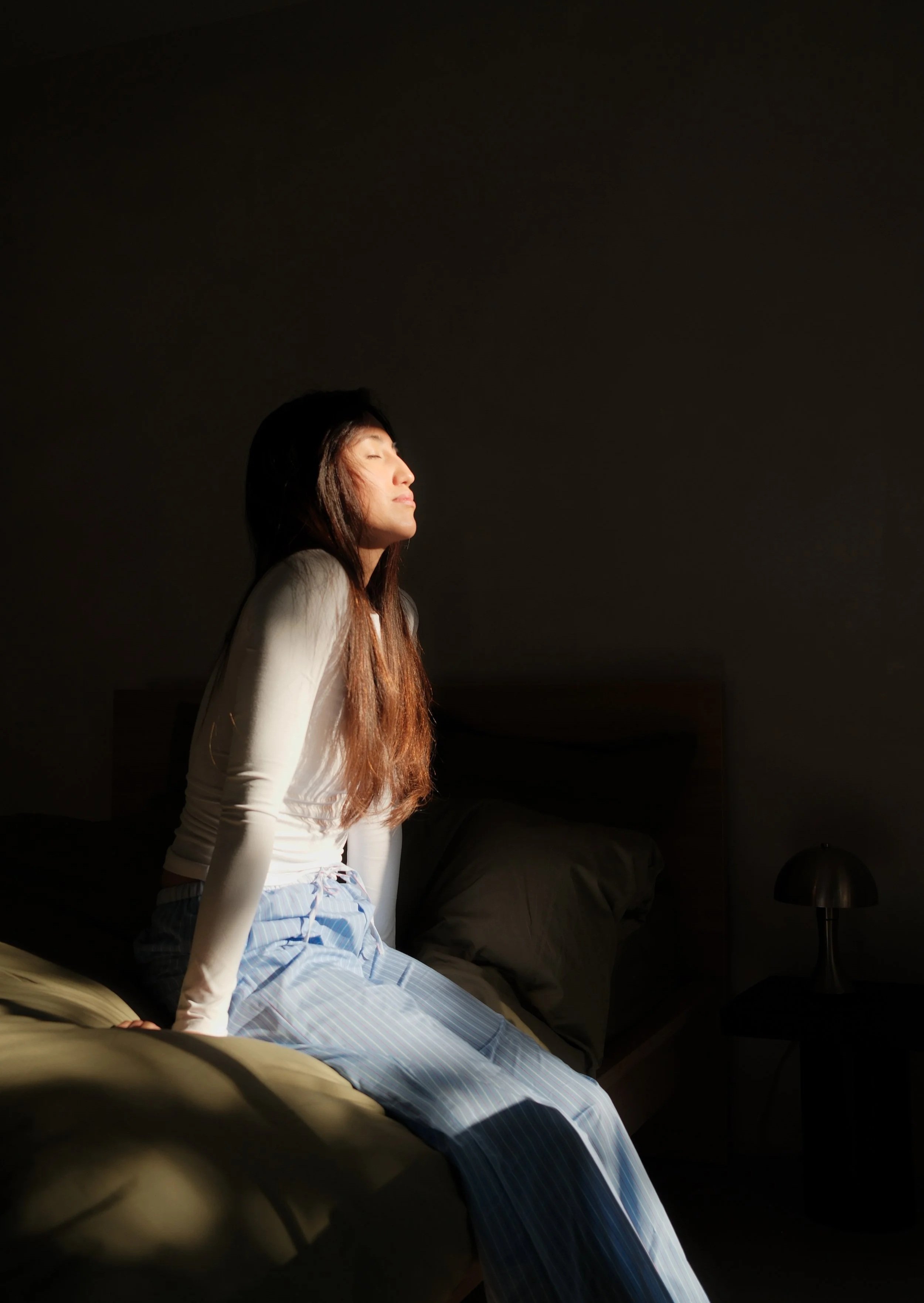 A woman with long brown hair sitting on a bed with her eyes closed and face tilted upward, illuminated by sunlight through a window, wearing a white long-sleeve shirt and light blue striped pajama pants, in a dimly lit room with a dark wall and a small table lamp on a bedside table.