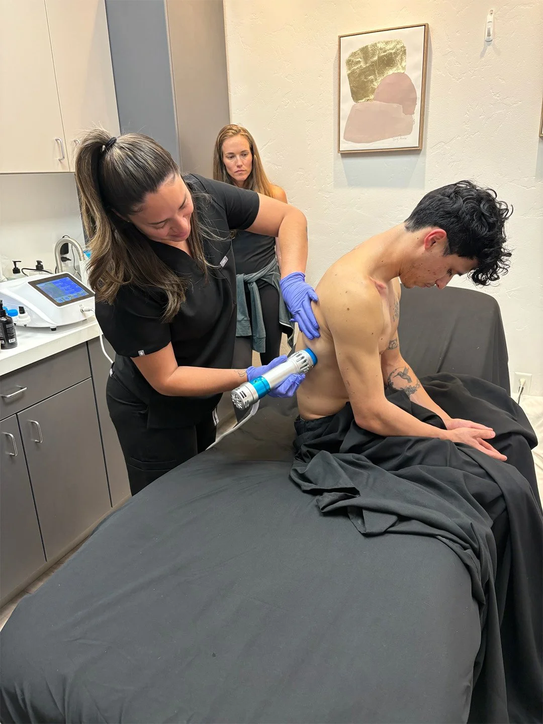 A woman in black scrubs uses a handheld device on a topless man sitting on a black-covered medical bed, while another woman observes in the background. Medical equipment and abstract wall art are visible in the room.