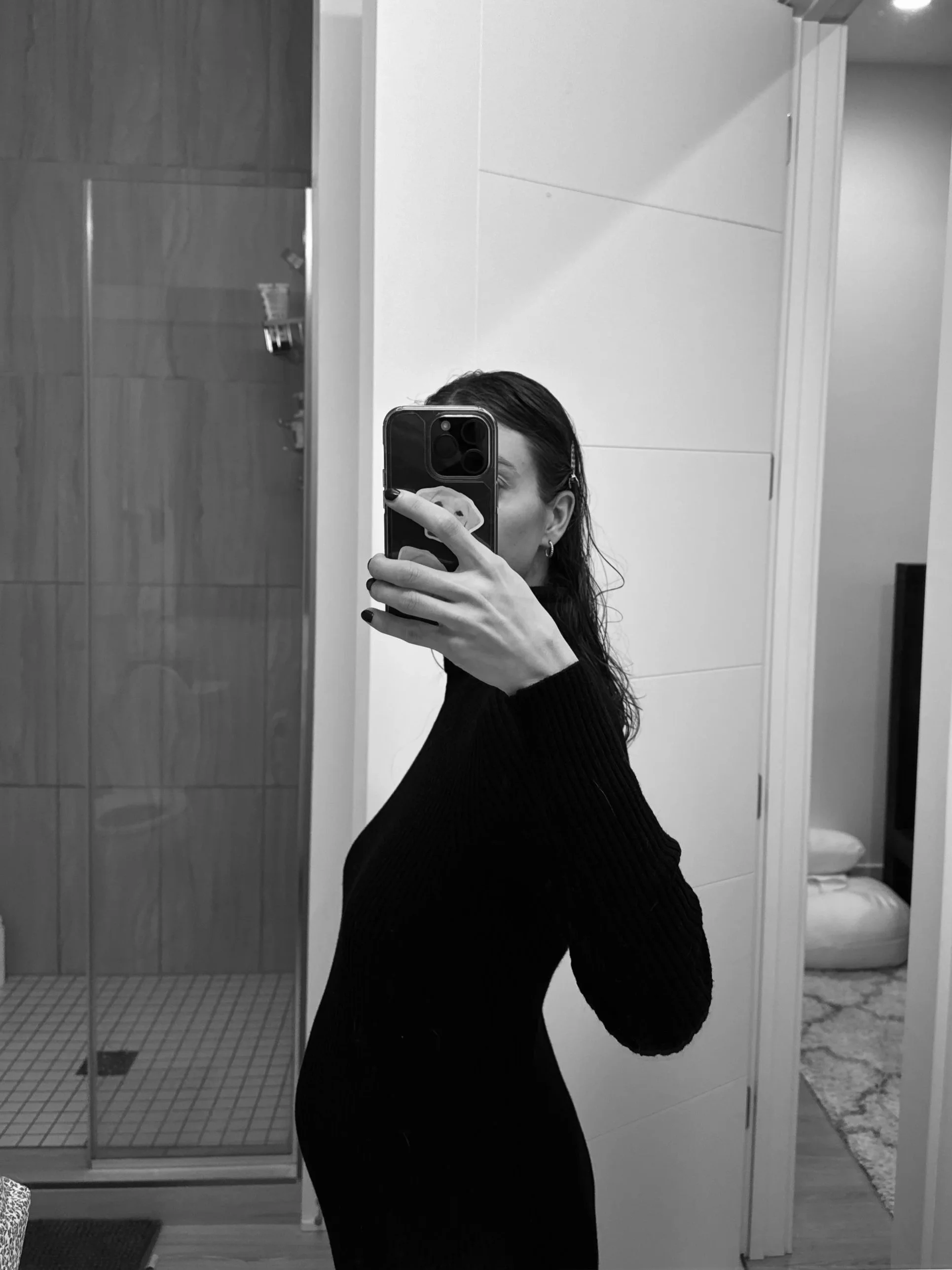 Black and white photo of a woman taking a selfie in a mirror at a bathroom or dressing room, wearing a long-sleeve black top, with visible earrings, and holding a smartphone with a patterned case.