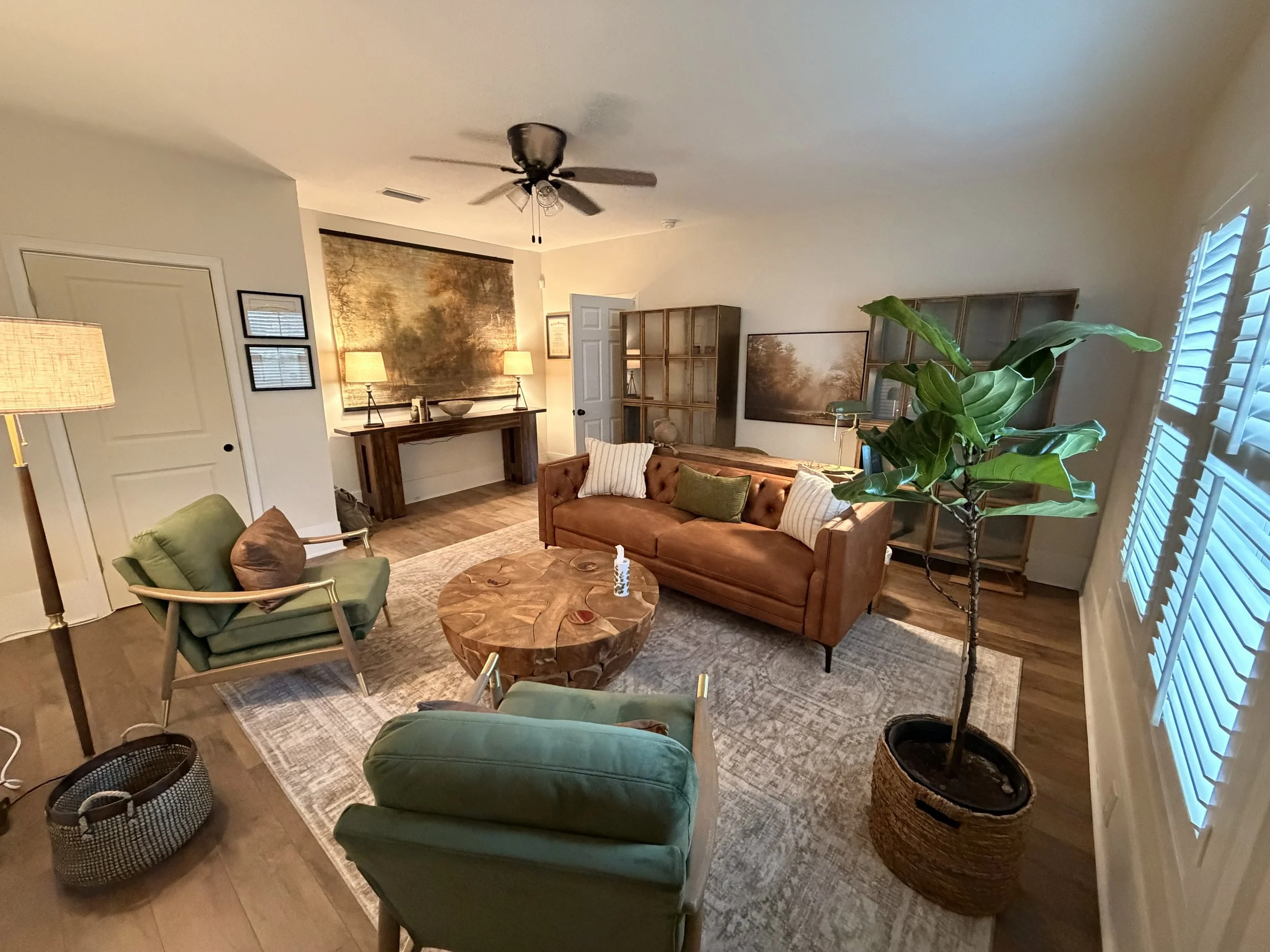 Living room with green armchair, brown sofa, large leafy plant, wooden coffee table, wall art, lamps, window with blinds, and wooden shelving units.