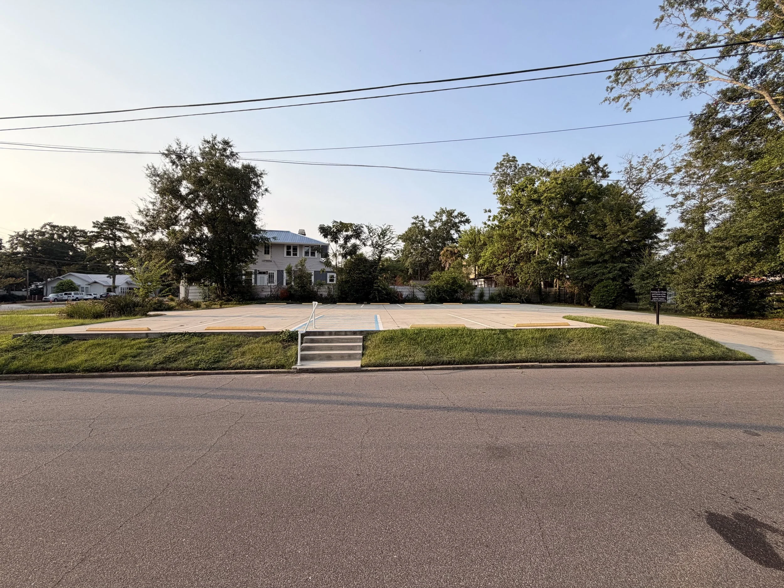 An empty parking lot with a small set of stairs leading up to a grassy area with trees and houses behind them, under a clear blue sky.