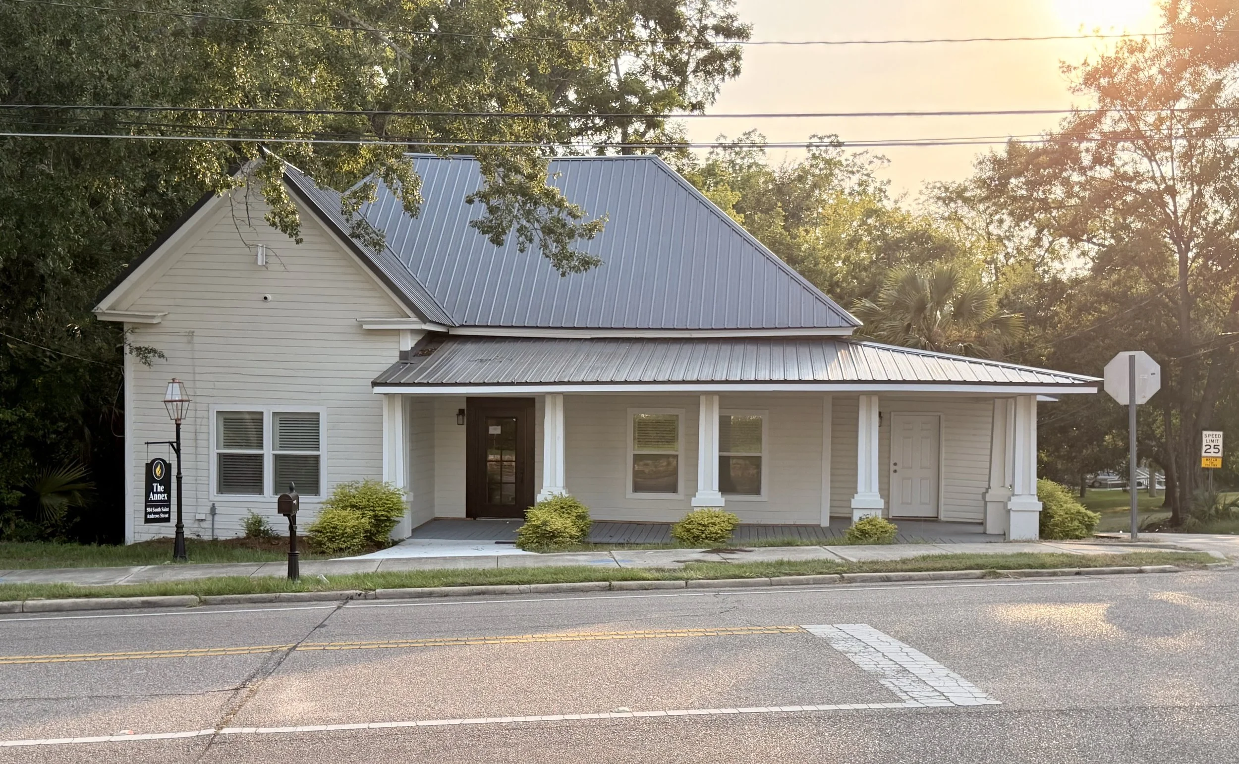White building with a metal roof and small porch, located beside a street with a speed limit sign, surrounded by trees and greenery, during sunset.