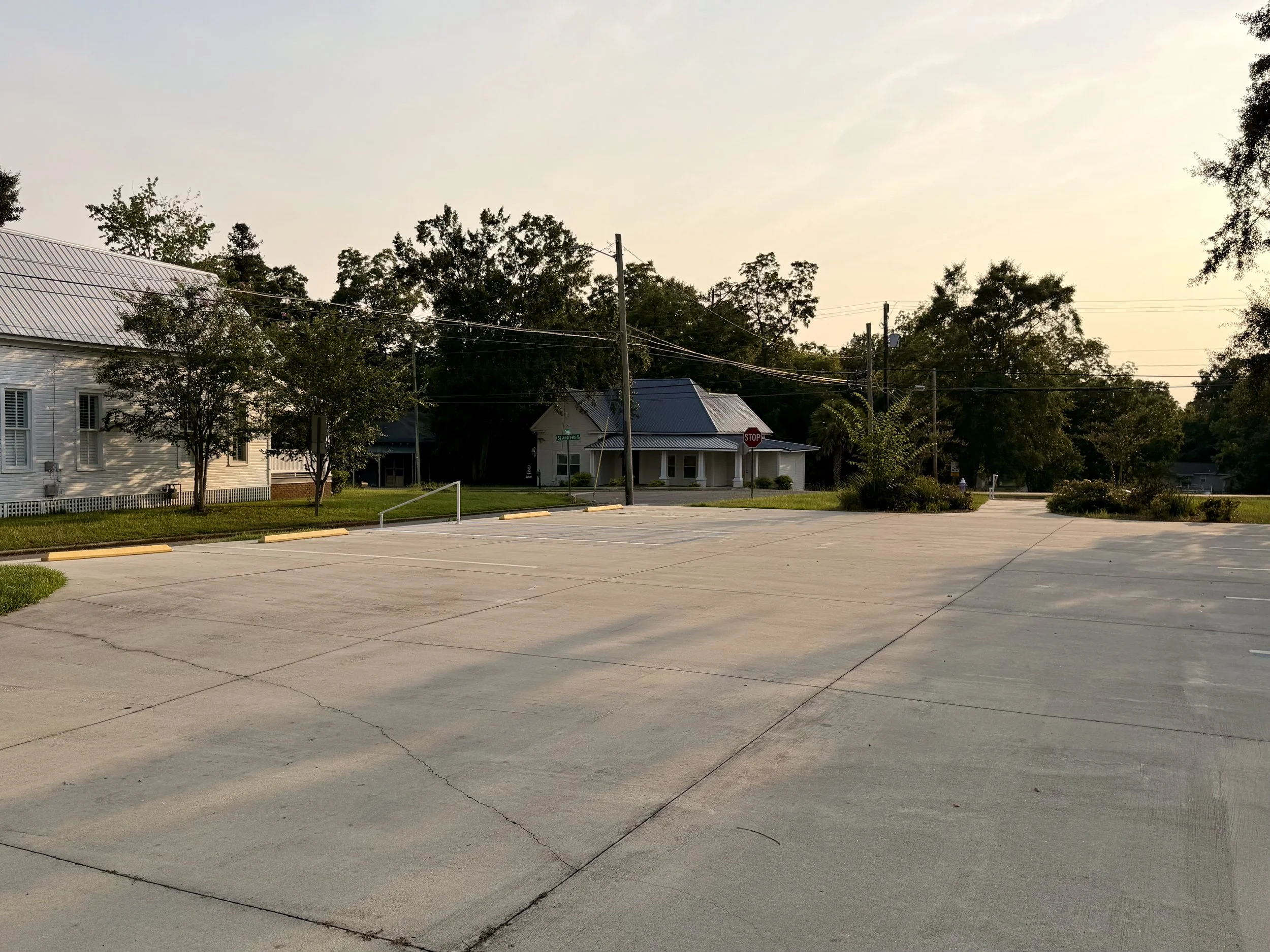 A parking lot with a few empty spaces, a house with a blue metal roof, trees, power lines, and a stop sign under a clear sky.