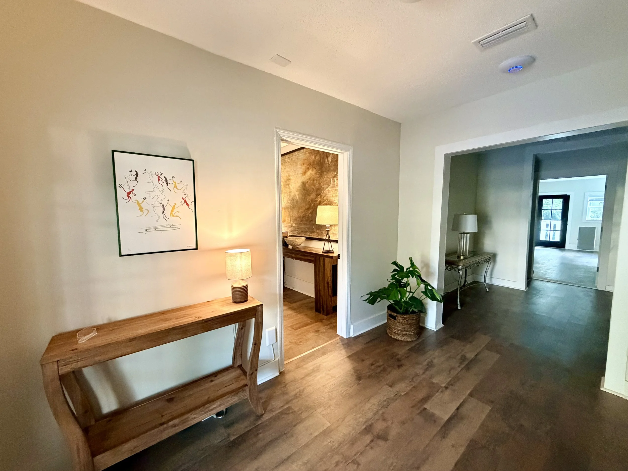 A living space with hardwood floors, a wooden console table with a lamp, artwork on the wall, a doorway leading to a room with a lamp, and a potted plant.