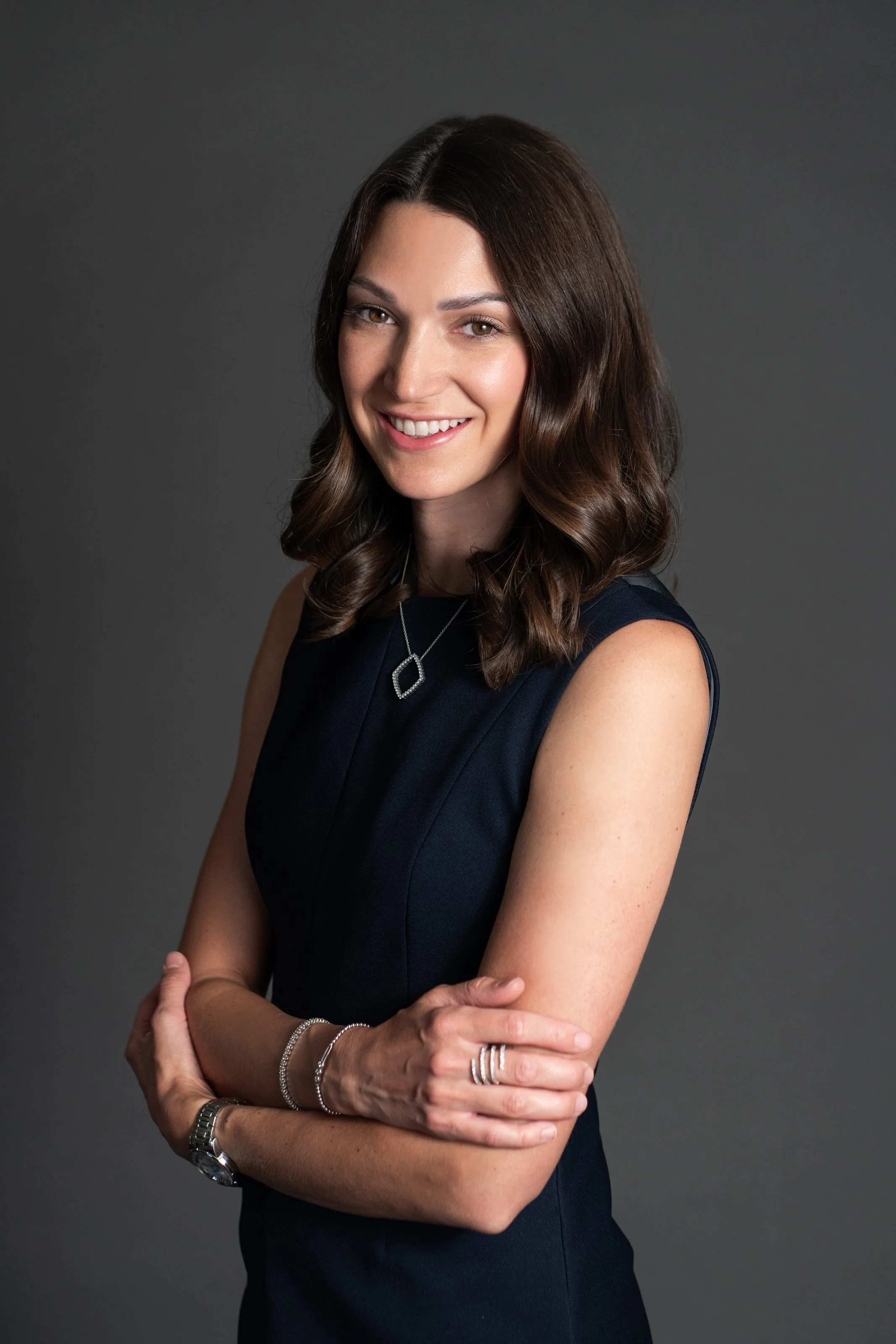A woman with brown wavy hair wearing a black sleeveless dress, silver jewelry, and a watch, standing with her arms crossed, smiling against a dark gray background.