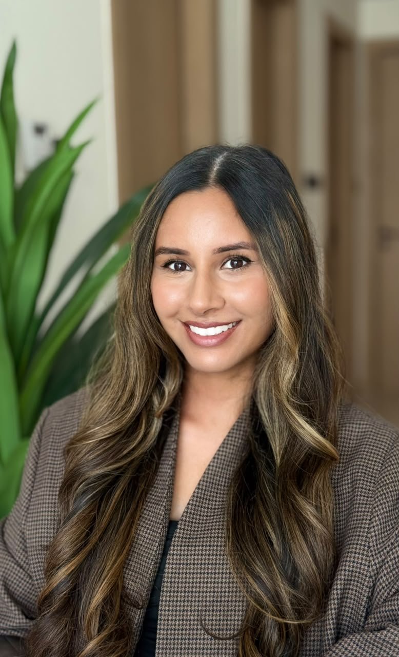 A psychotherapist in Dubai with long, wavy brown hair smiling at the camera, wearing a brown checkered blazer in an indoor setting with a green plant in the background