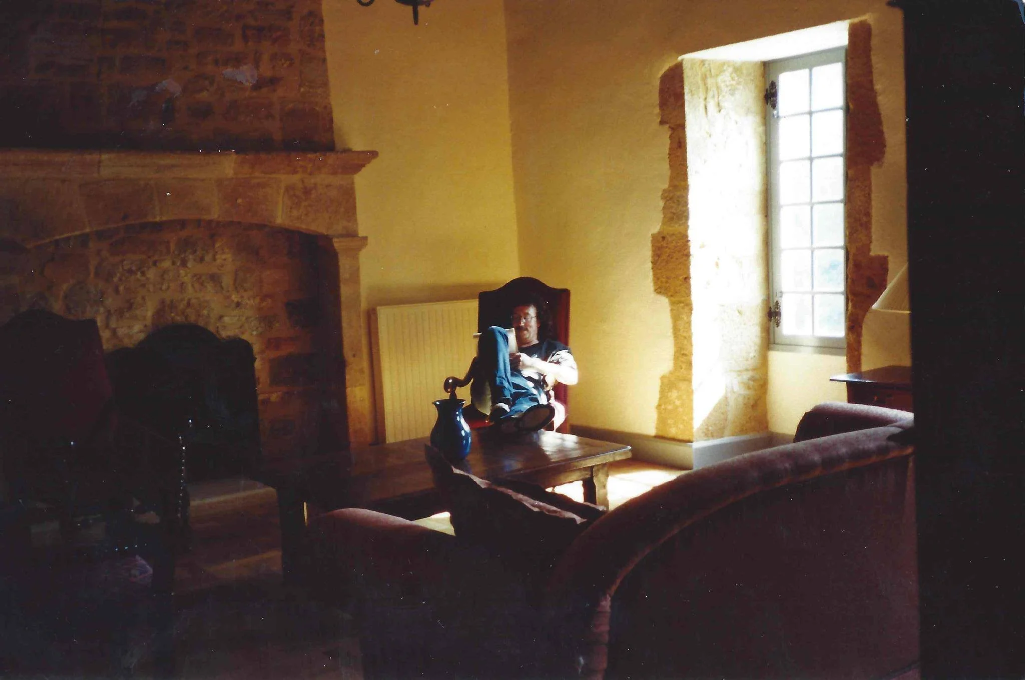 A man with glasses sitting in a high-back chair in a room with exposed stone wall and a large window allowing sunlight to enter. A wooden coffee table with a blue vase is in front of him, and a pair of shoes are on the floor nearby.