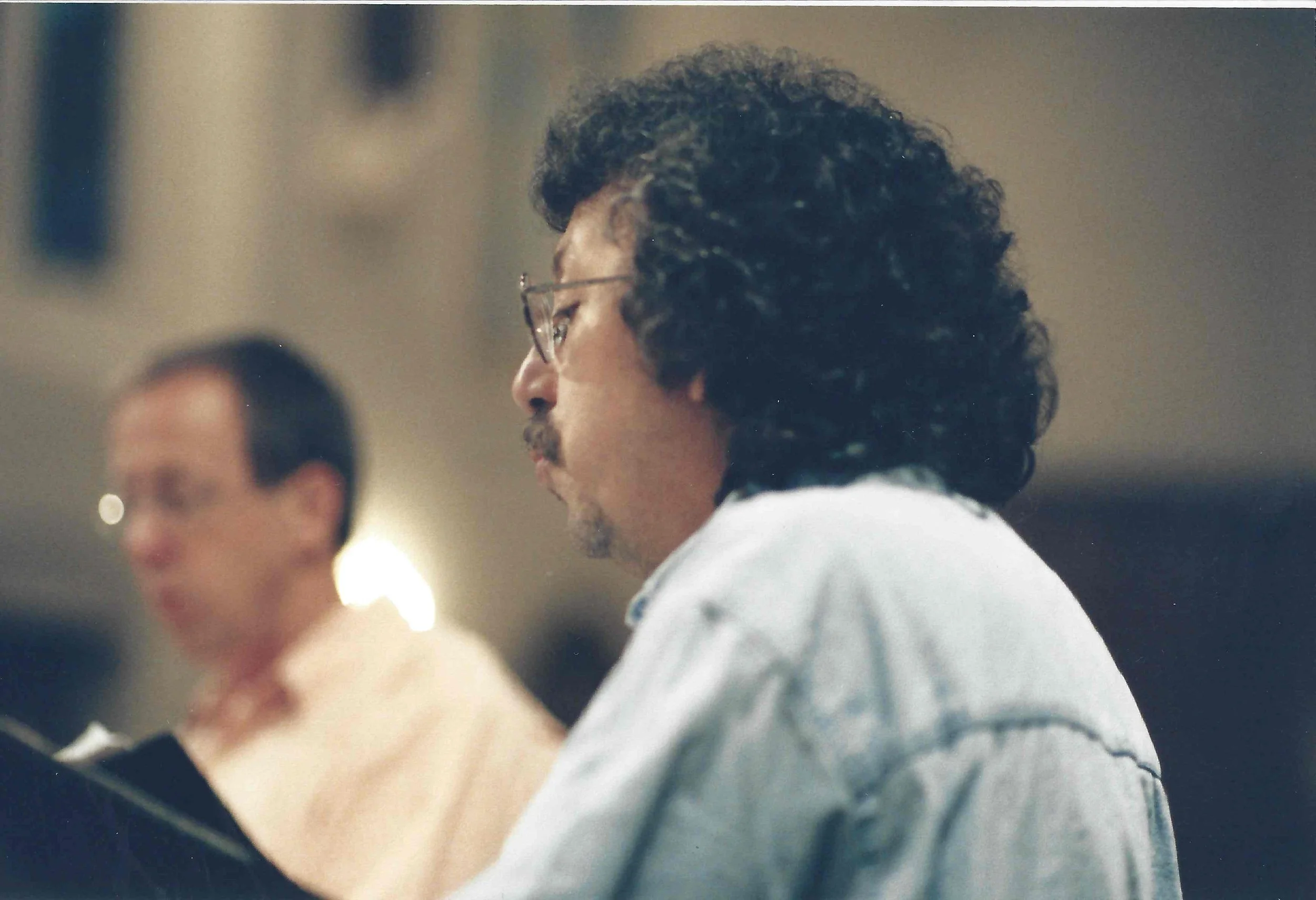 A person with curly dark hair and glasses, wearing a light-colored shirt, looking at a computer or other device, with another person blurred in the background.