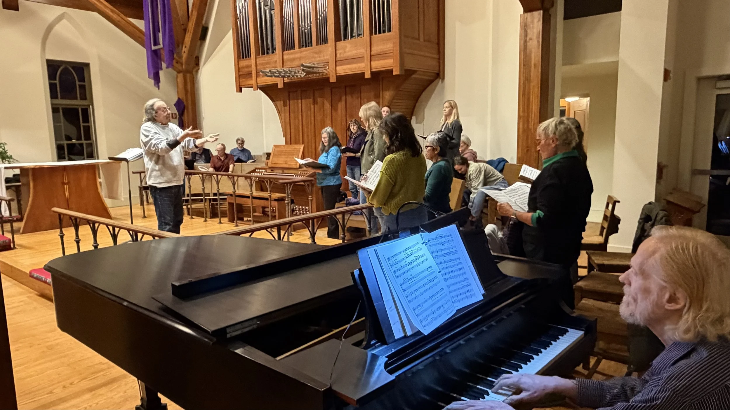 A group of people gathered in a church or concert hall, participating in a choir rehearsal or singing session, with a pianist playing a digital piano in the foreground.