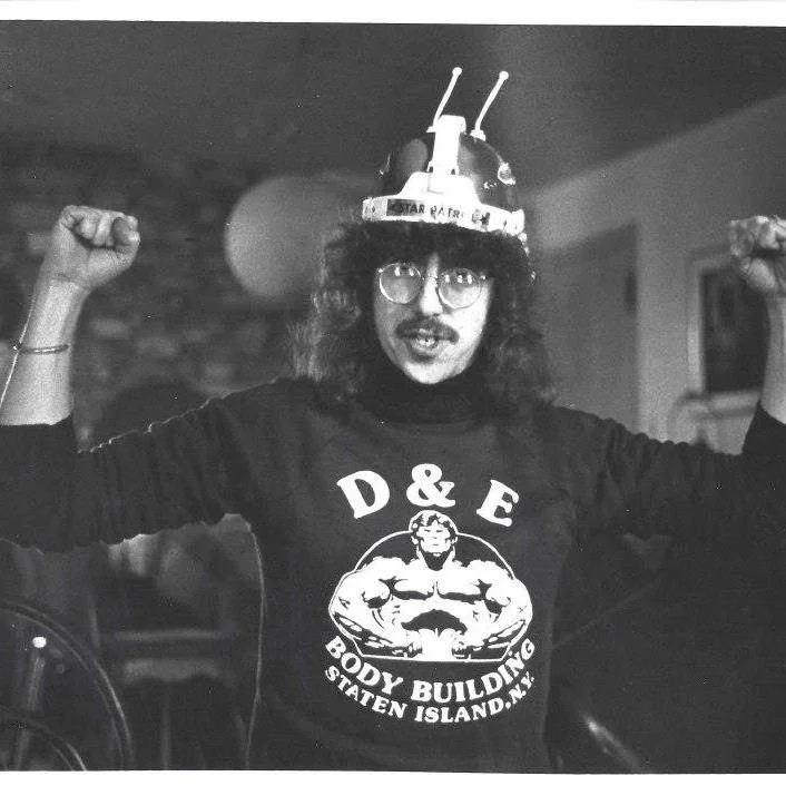 A young person with long hair, glasses, and a mustache wearing a science fiction hat that looks like a robot or spaceship. They are flexing their muscles and wearing a sweatshirt that says 'D&E Body Building Staten Island N.Y.' with a muscular cartoo