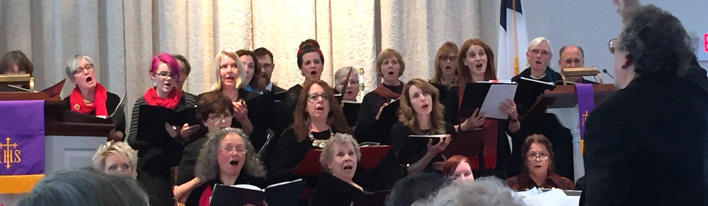 A choir of men and women singing with a conductor leading them in a church or religious setting.
