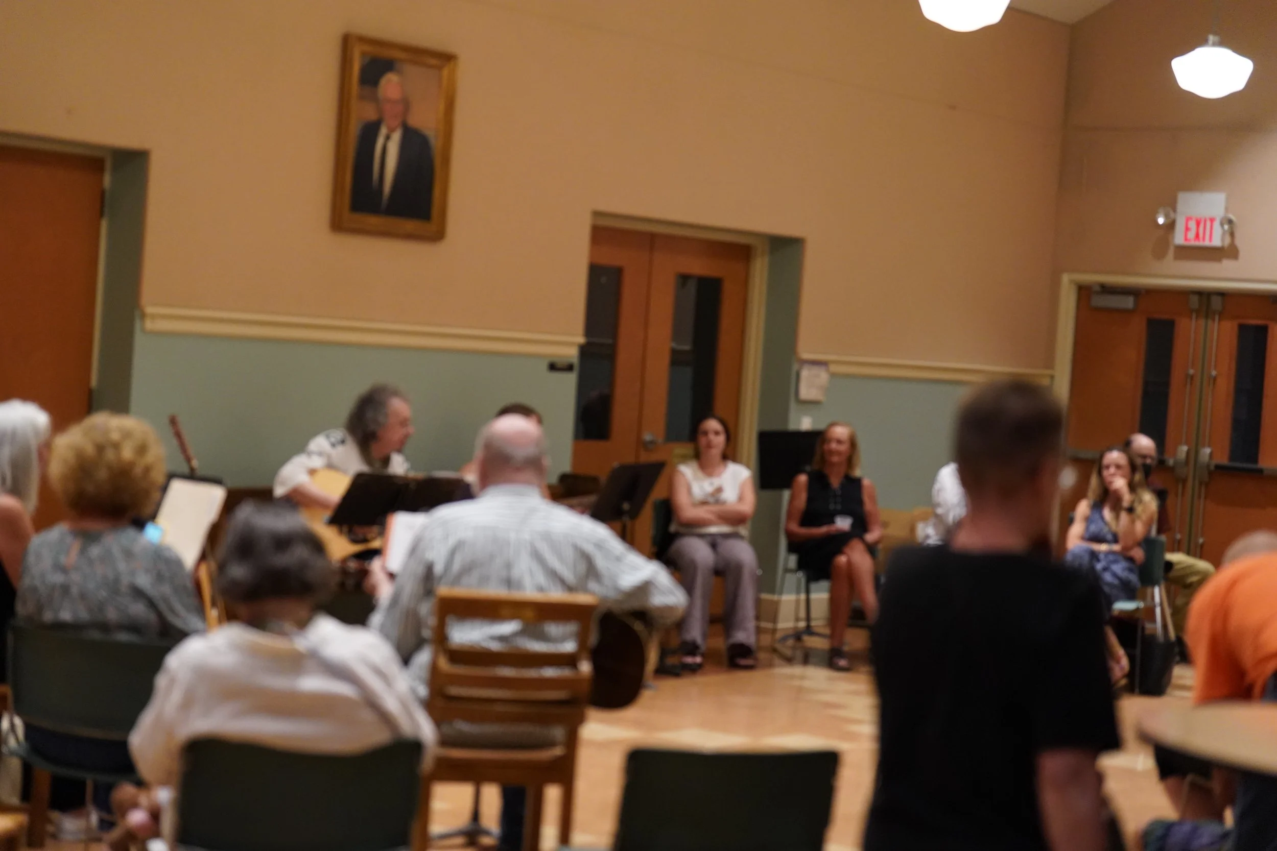 A group of people sitting in a room, some holding musical instruments, with a woman in the center speaking or singing, in a room with warm-colored walls, framed pictures, and hanging light fixtures.
