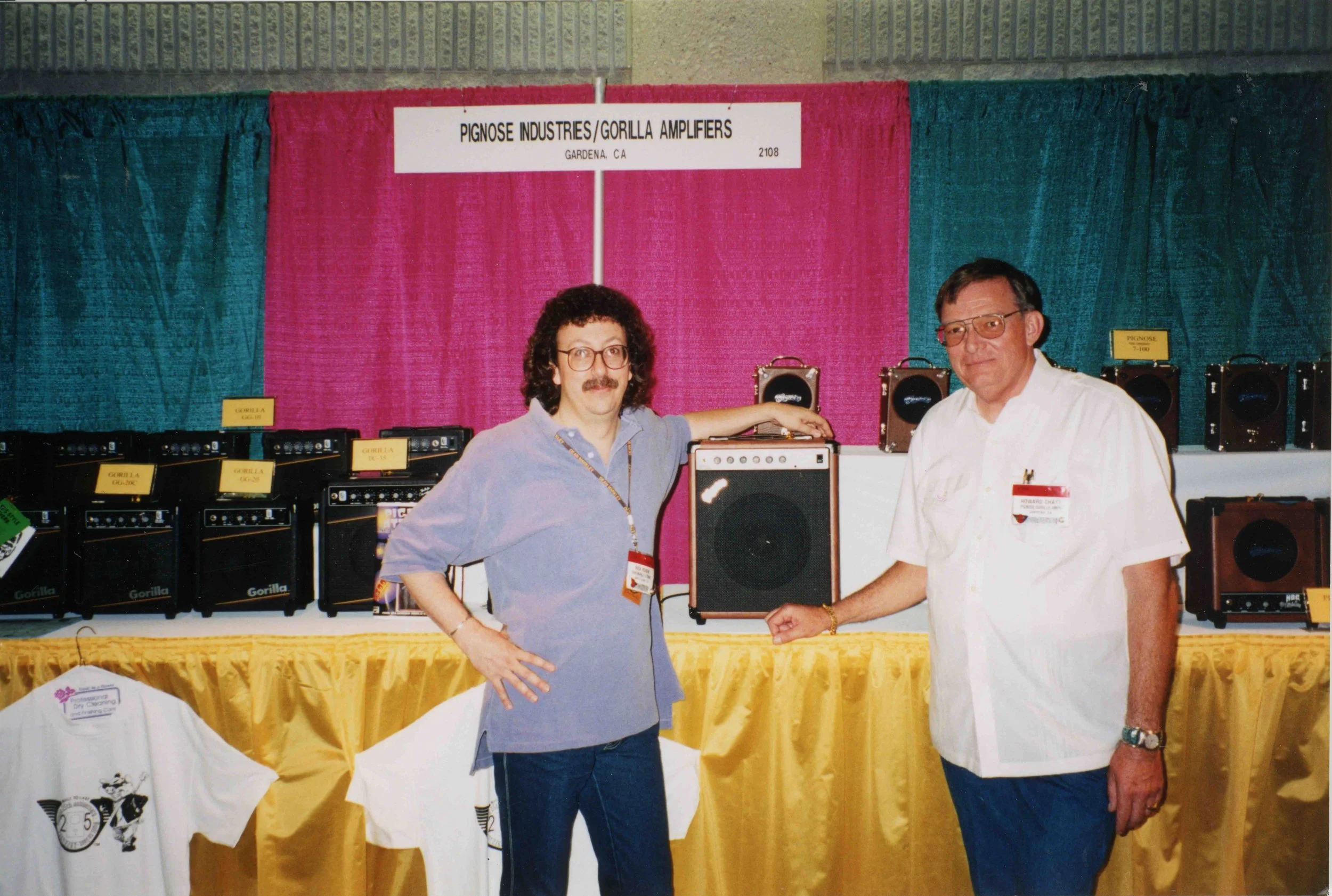 Two men standing in front of a display of Gorilla brand amplifiers at a tradeshow or exhibition, with a colorful background and yellow tablecloth.