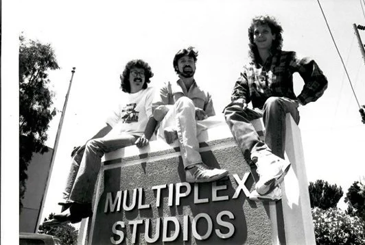 Three young men sitting on top of a sign that reads 'MULTIPLEX STUDIOS,' with trees and utility poles in the background.