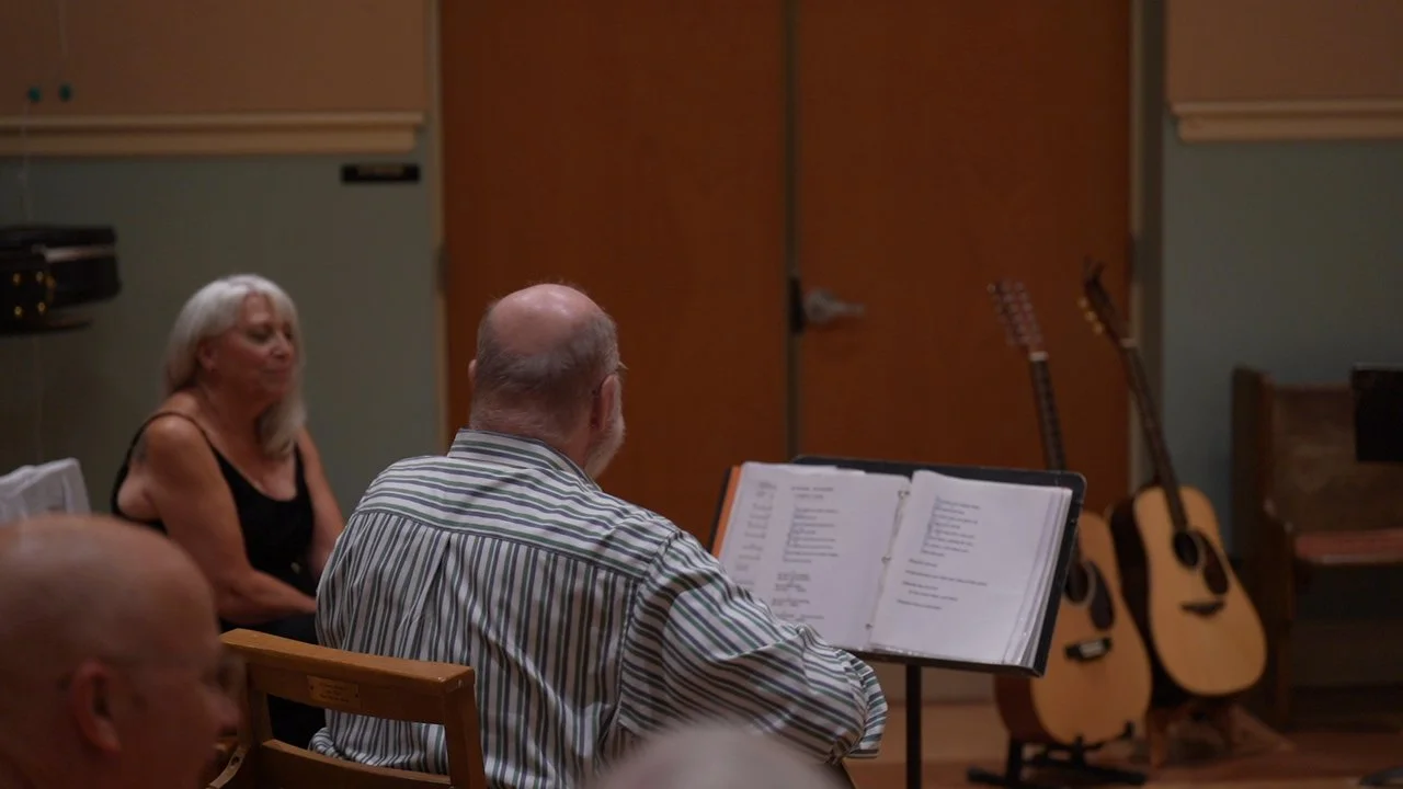 Older man with glasses and striped shirt reading music lyrics, woman with gray hair and black dress sitting nearby, two guitars leaning against the wall in a music practice room.