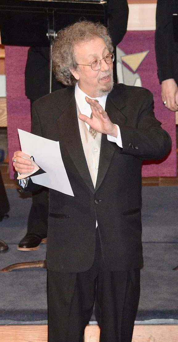 A man with curly gray hair, glasses, and a beard wearing a black suit and white shirt, holding a piece of paper in his left hand while making a gesture with his right hand, standing indoors at an event or gathering.