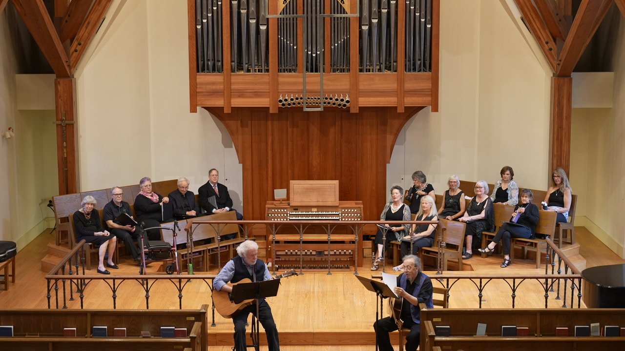 A group of people in black clothing sitting on benches and chairs on a wooden stage in a church or concert hall, with two men playing guitars in front of the group, and a large organ in the background.