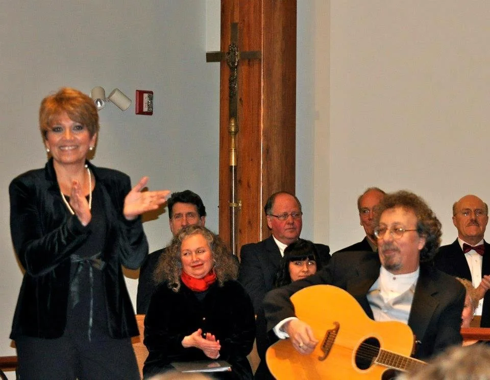 Woman clapping and smiling at a gathering with people seated, including a man playing a guitar, in a room with a cross on the wall.