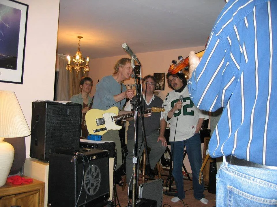 A group of five people in a living room with musical instruments and microphones, appearing to perform or practice music.