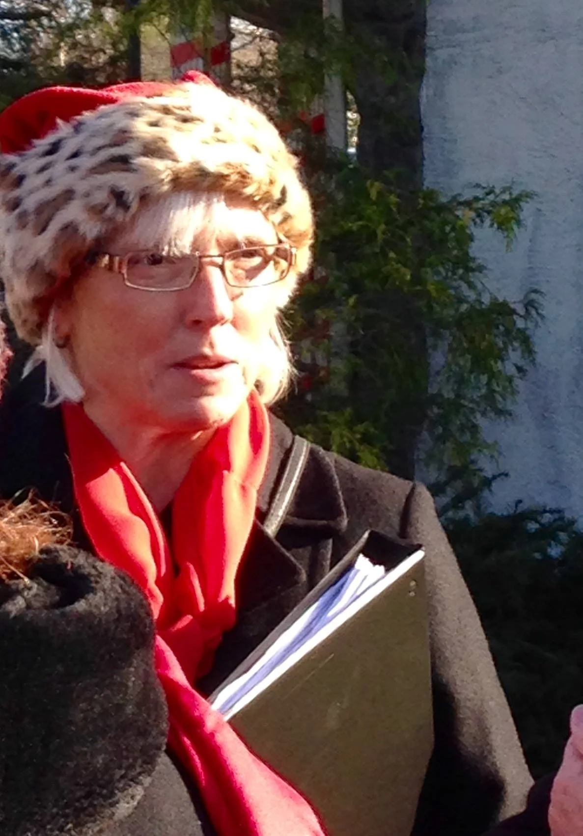 A woman wearing glasses, a leopard print hat, a red scarf, and a black jacket, holding a notepad outdoors near water with trees in the background.