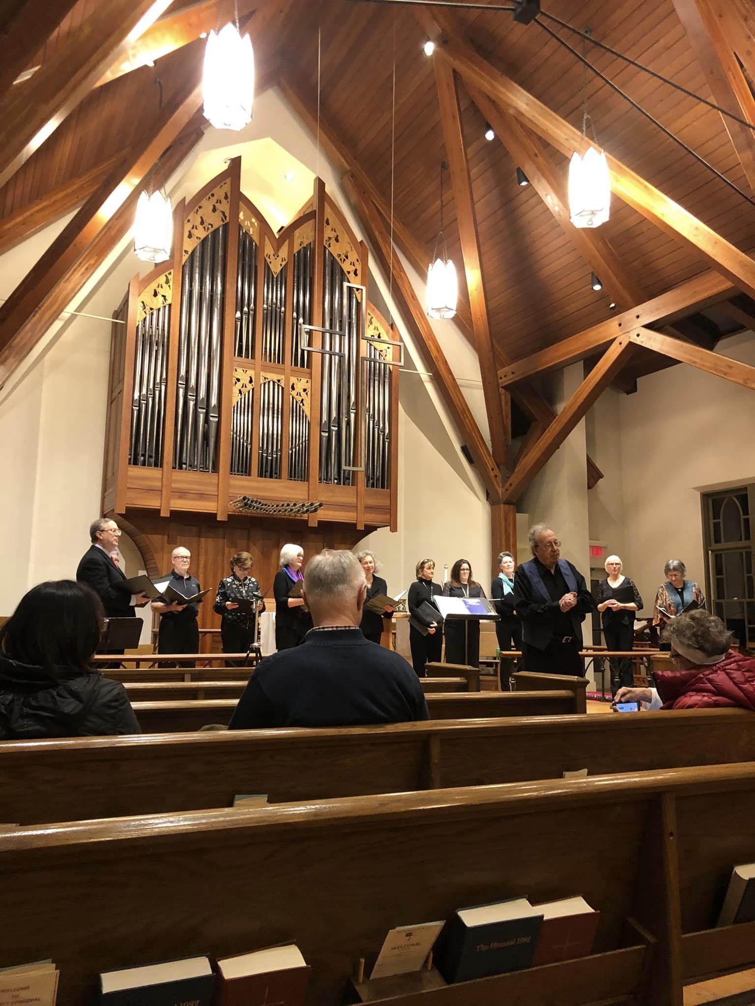 A choir sings in a church with a large pipe organ and wooden ceiling, while an audience watches in pews.