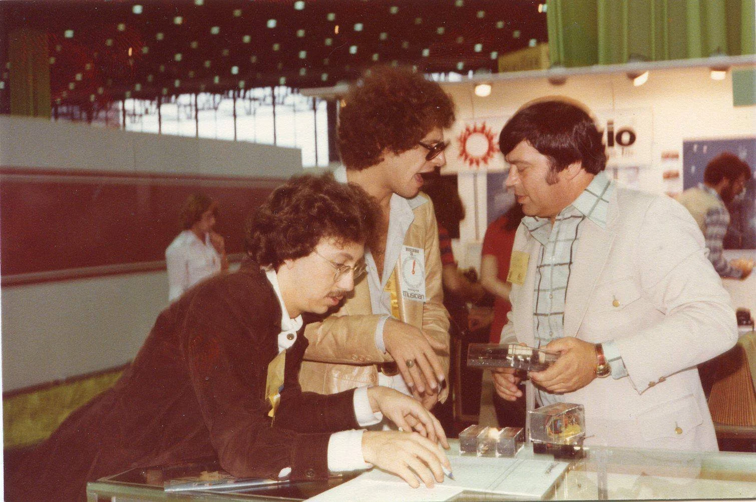 Three people with 1970s hairstyles and clothing examine items on a display table at an indoor event.