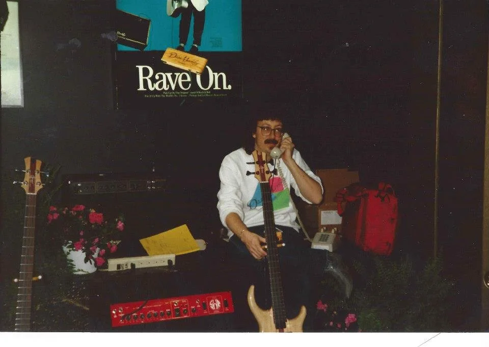 A man with glasses, a mustache, and curly hair sitting on a black bench, holding a landline phone, with a guitar strap around his neck. There is an acoustic guitar leaning against the bench, a red effects pedal on the floor, and pink flowers in a whi