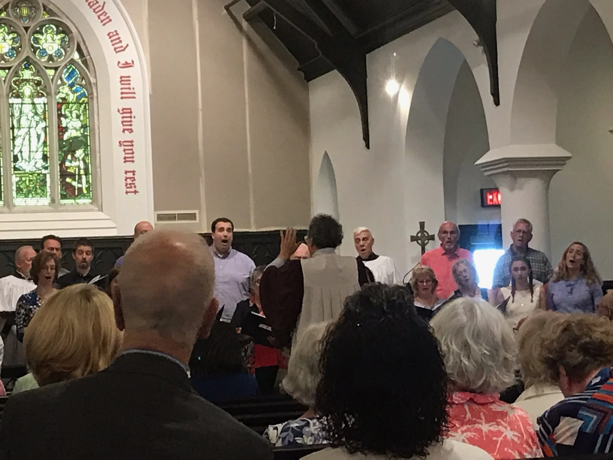 A choir performs inside a church with stained glass windows and white walls. The congregation watches, with some people singing and others taking photos. The church has high ceilings, stone columns, and a religious cross on the wall.