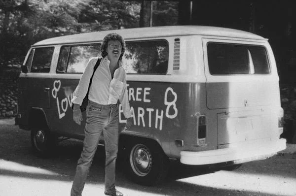 A woman with curly hair, wearing glasses and casual clothing, standing in front of a van with the words "Free Earth" and hearts painted on it.