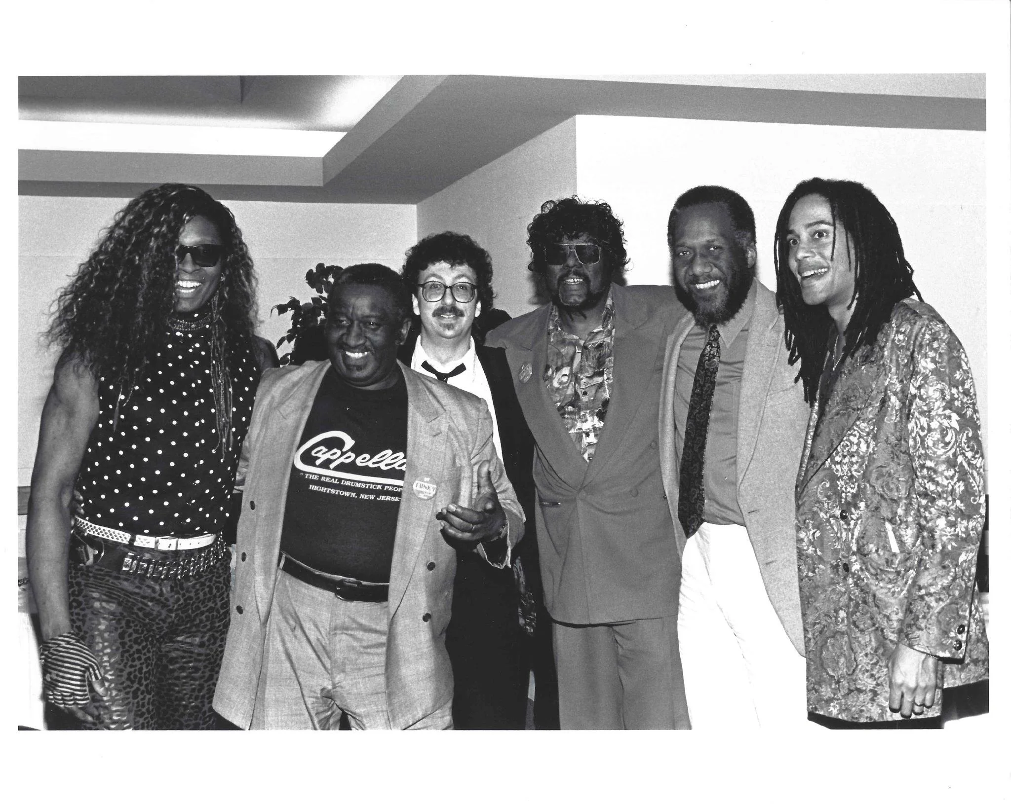 Six people standing together at a social gathering. Some are smiling and dressed in colorful clothing, with one person wearing sunglasses and a jacket, and others in patterned outfits and suits.