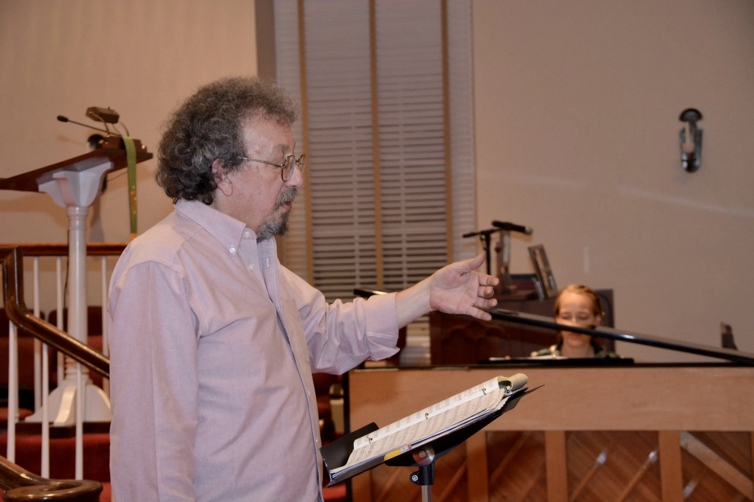 A man with curly hair, glasses, and a beard wearing a light pink shirt is conducting or singing, holding a sheet music with a music stand in front of him. A woman with glasses and a dark top is playing the piano in the background. The setting appears
