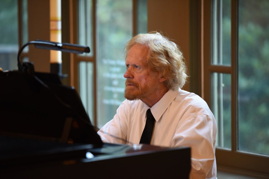 An elderly man with curly blond hair and a beard, wearing a white shirt and black tie, sitting at a piano near large windows with a view of trees outside.