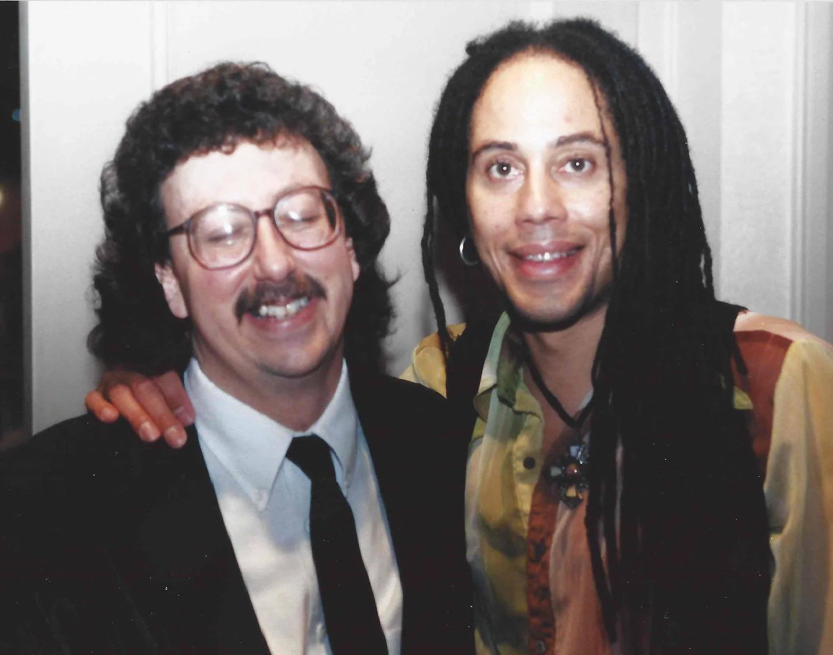 Two men smiling and posing closely together for a photo. The man on the left has curly dark hair, glasses, and is wearing a tuxedo with a bow tie. The man on the right has dreadlocks, is wearing a colorful patterned shirt, and has his arm around the 