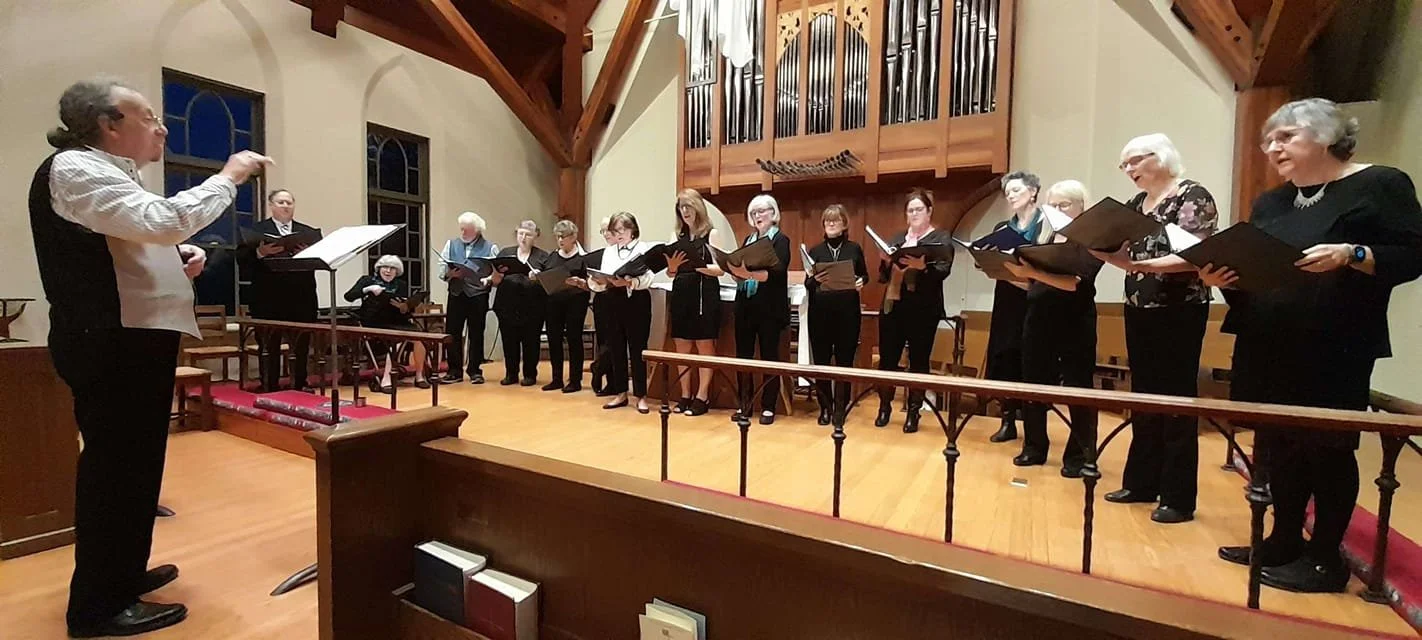 A choir practicing in a church. The conductor stands in front, guiding the group of singers, who hold music folders. The setting features wooden beams and an organ pipe display in the background.