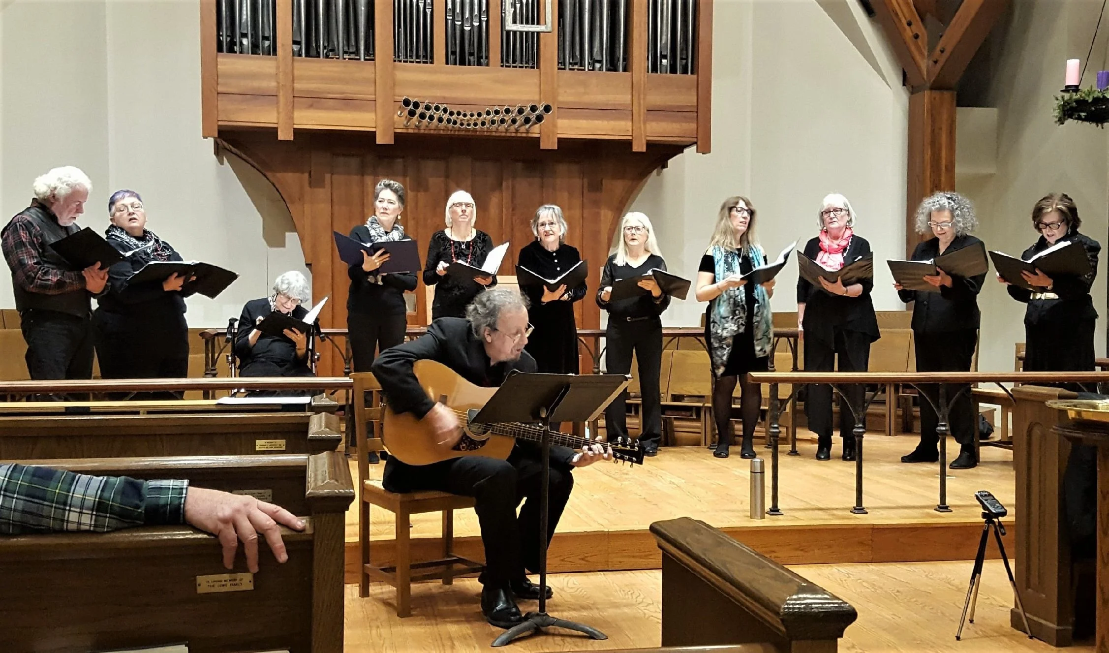 A choir of eleven older women and one man playing guitar, performing in a church or concert hall. The women are holding black folders, singing, while the man is seated, playing an acoustic guitar. The background features a large wooden organ pipe str
