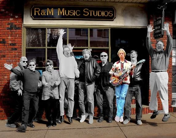 A group of nine people, mostly men, standing in front of R&M Music Studios, with some raising their hands and one person holding a guitar. The building has a brick exterior and large windows.