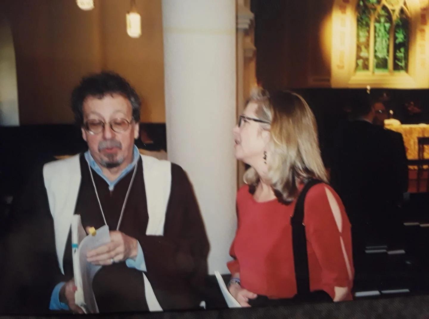 A man with glasses and a beard holding papers and a woman with glasses and earrings in conversation inside a warm, dimly lit restaurant or cafe.