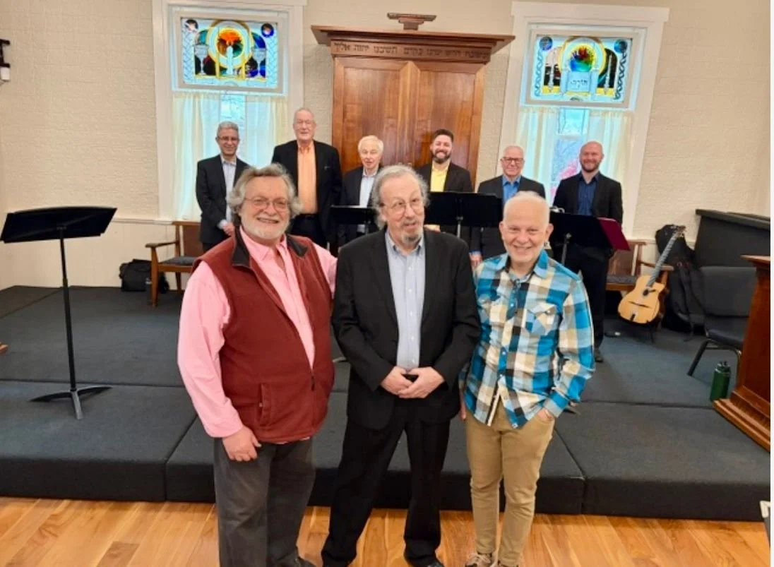 A group of nine men posing inside a church with stained glass windows behind them, some standing on a stage with musical instruments, while three men stand in the foreground.